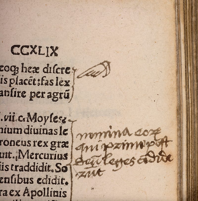 Detail of page from an old book with black text. A hand-drawn manicule with sleeve is pointing towards a passage. Beneath it there are some hand-written notes. The passage has also been highlighted with a hand-drawn bracket.