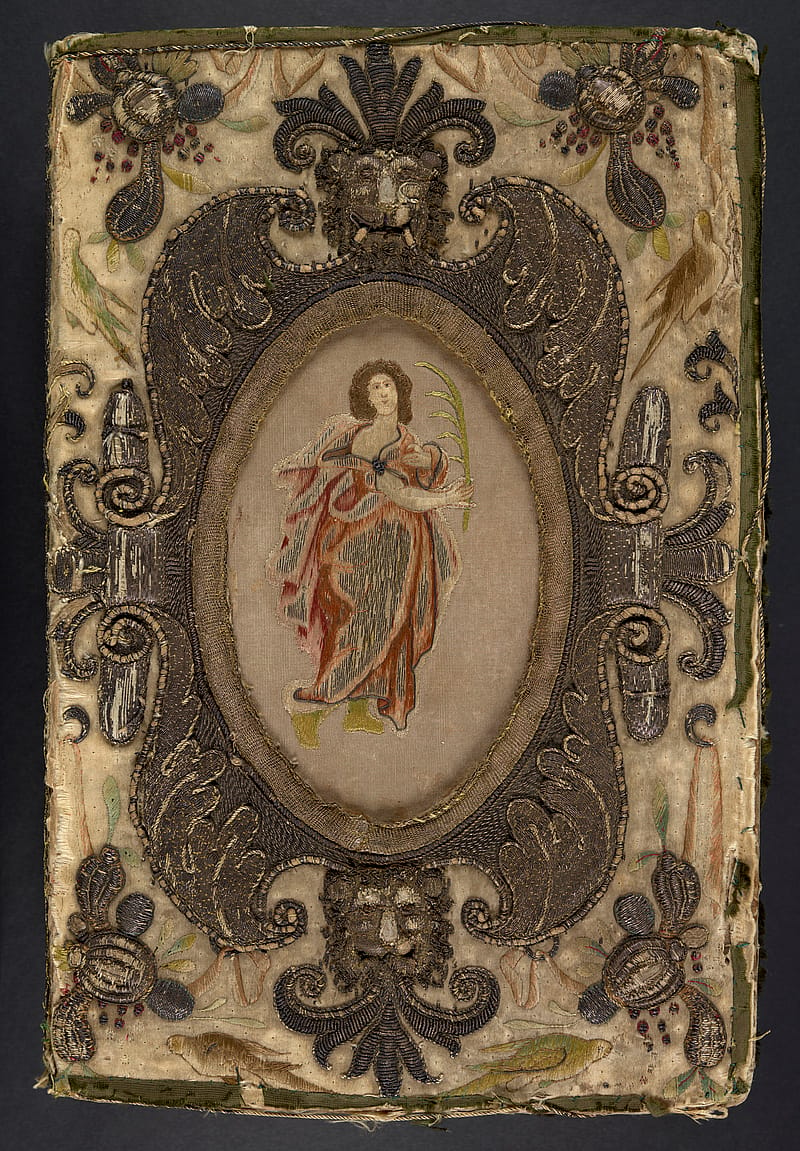 Front cover of an old book with embroidered binding. The binding depicts a full-length female figure within a cartouche. She has brown hair and is wearing a red and white flowy dress. She is holding a large feather. Lion heads decorate the top and bottom of the cartouche. Around the cartouche are decorative embroidered floral designs.