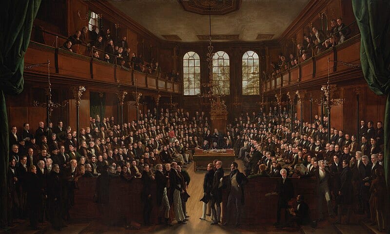Painting of the interior of The House of Commons. There are hundreds of male figures in dark suits seated either side of a central table at which sits a man in a wooden, carved chair with a high back.